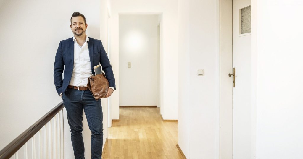 Tim Post steht mit einer Ledertasche unter dem Arm im Obergeschoss-Flur eines Wohnhauses mit Holzboden.