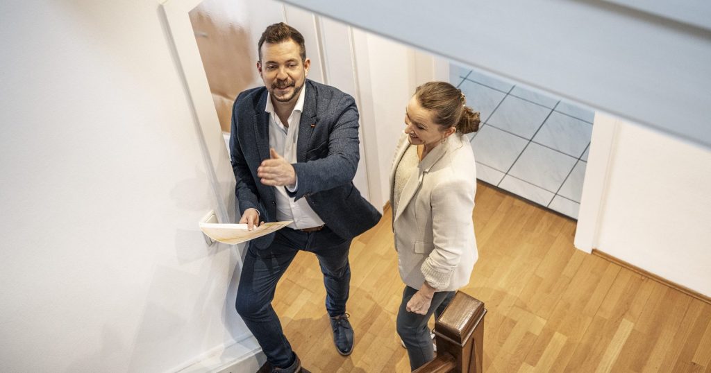 Tim Post geht mit Unterlagen in der Hand eine Holztreppe hinauf und erklärt einer Frau im hellen Blazer das Obergeschoss eines Wohnhauses.